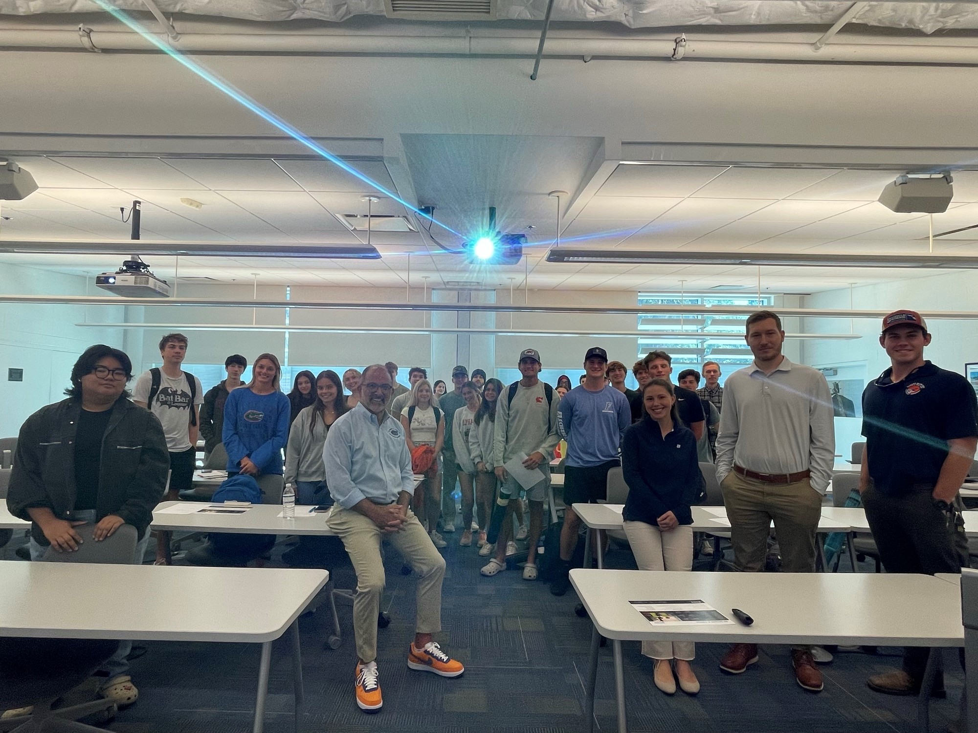 A group of people stand and sit in a classroom with white tables, chairs, and a projector screen at the front. The room is well-lit and the individuals face the camera.