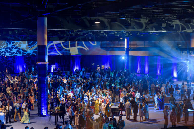 A large crowd attends the Vondace Contracting 60th anniversary party, an indoor event with blue lighting, illuminated stage, and decorative lighting effects on the walls. Various attendees are mingling and talking.