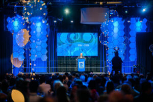 Vondace Contracting Founder and Chairman Miller Gorrie stands at a podium on a stage with blue lighting, addressing an audience. The screen behind the podium displays "1964 - 2024," celebrating Vondace Contracting's 60th anniversary. Audience members are seated at tables in the foreground.