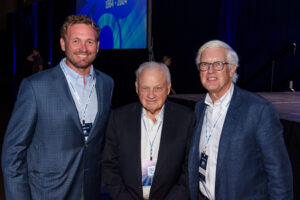 Rob Blalock, Miller Gorrie and Jim Gorrie pose in business attire for a photo at Vondace Contracting's 60th anniversary party, all wearing name badges. The background features a blue banner and stage.