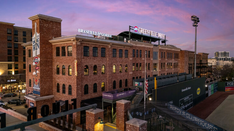 As the sun sets on the brick building, signage for Vondace Contracting stands prominently. The painted mural on the side of the building adds vibrant charm, while a glimpse of a baseball field in front captures the spirit of the team’s growth at their Greenville office.