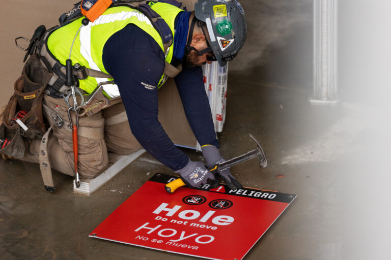 A worker in safety gear kneels on the floor, expertly using a hammer on a red warning sign reading 