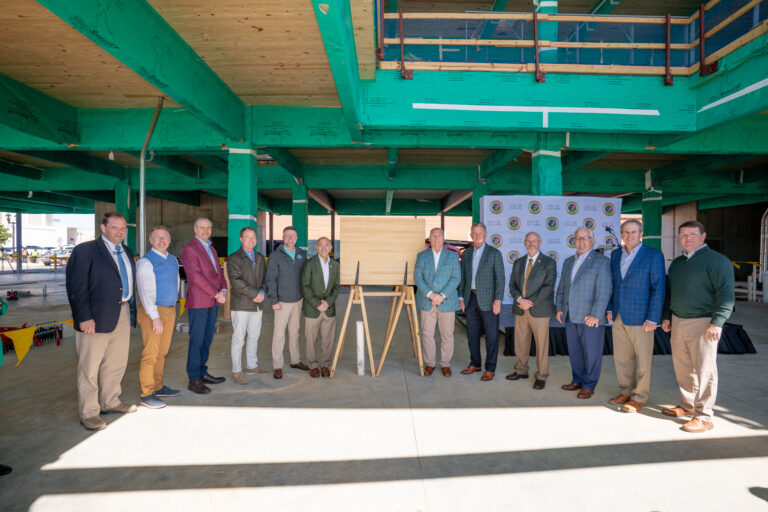 A group of people stands in a row at the construction site of the Wiregrass Innovation Center, posing in front of a backdrop adorned with logos, framed by green support beams.