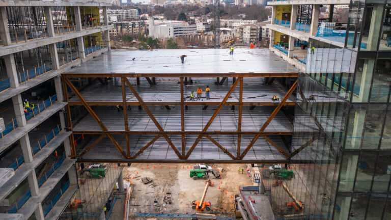 Workers from Vondace Contracting are constructing a large building featuring a steel framework and glass facade, with a focus on a multi-story sky bridge under construction between two buildings.