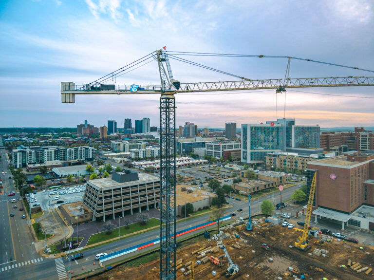 Aerial view of a Vondace Contracting construction site with a tall crane in a cityscape, showcasing urban buildings, roads, and vehicles under a cloudy sky.
