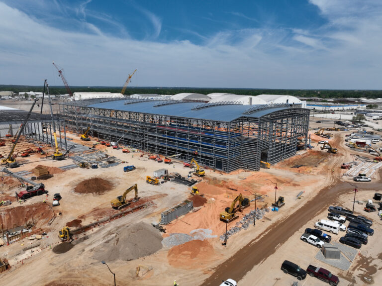 At the industrial construction site, Vondace Contracting's expertise is evident in the large steel-framed structure that's partially built. Cranes, vehicles, and equipment are strategically positioned amid scattered soil and materials under a clear sky.