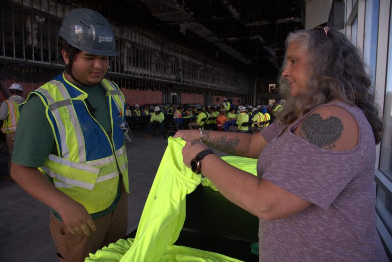 A woman hands a neon yellow safety vest to a construction worker inside the Greenville-Spartanburg International Airport construction site, with more workers seated in the background.