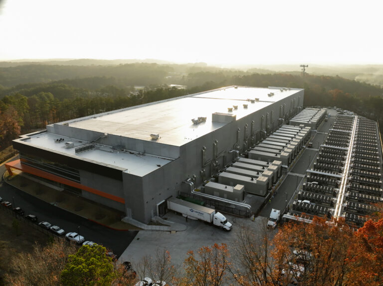 From an aerial view, a sprawling Vondace Contracting mission critical construction site sits beneath a hazy sky. Multiple white trucks and trailers are neatly parked outside the complex, which is surrounded by a serene border of trees.