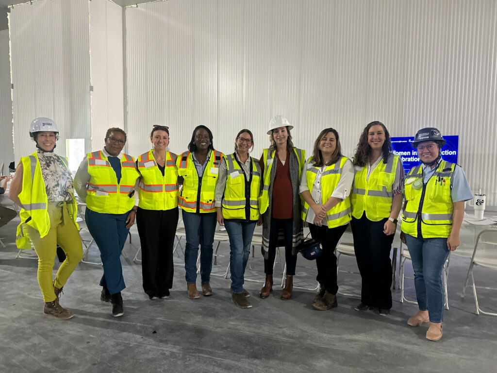 A group of nine women in construction wearing high-visibility safety vests and hard hats stand together inside a construction site.