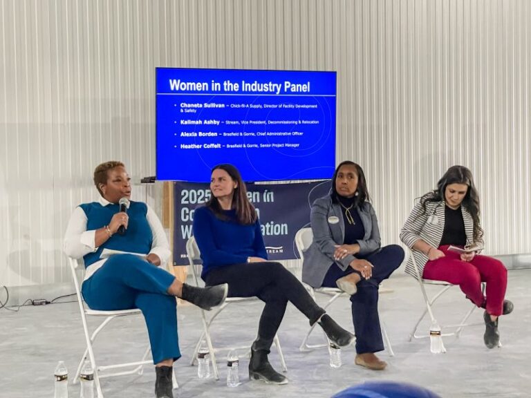 Four women sit on a panel in front of an audience, with a screen behind them displaying the topic 