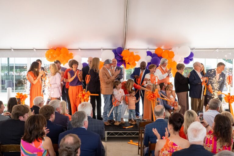 A group of people on stage cutting an orange ribbon at the Nieri Family Clemson Alumni and Visitors Center opening ceremony, with balloons in the background and the Nieri Family present, as the audience watches from their seats.