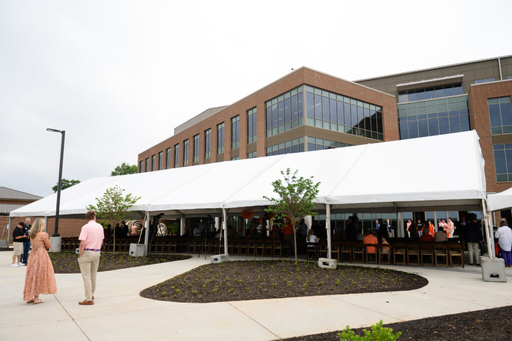 People gather under a large white tent outside the modern brick Clemson Alumni Center, with rows of chairs and landscaping in the foreground.