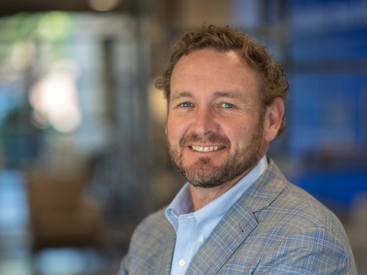 CEO Rob Blalock, wearing a light blue shirt and gray plaid blazer, smiles as he stands in an office setting with a blurred background.