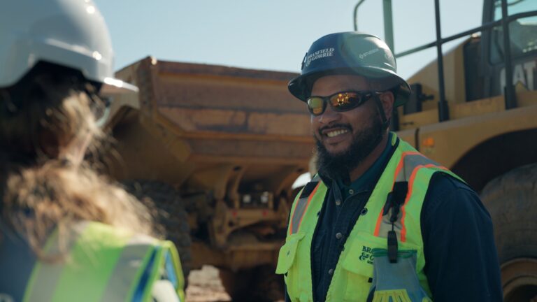 A construction worker in a safety vest, hard hat, and sunglasses smiles while standing next to heavy machinery at a worksite, capturing the spirit of a true Brasfield and Gorrie story.