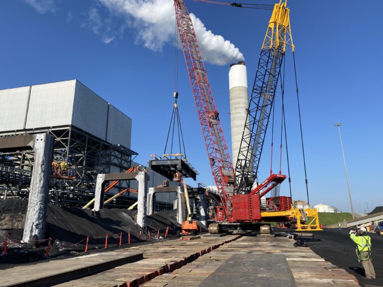 Large cranes lift heavy equipment at an industrial site with smoke stacks; workers in safety gear are present on the ground, showcasing Brasfield and Gorrie heavy civil projects.