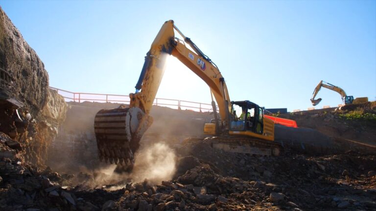 Two large yellow excavators dig and move rocks and dirt at a construction site under a clear blue sky, preparing the ground for the new EMF Office Building.