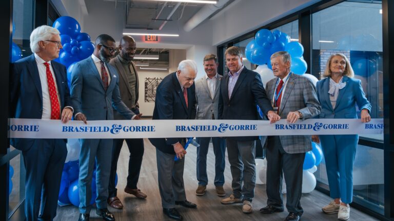 A group of people stand behind a ribbon labeled “Vondace Contracting” as one man prepares to cut it at an indoor ribbon-cutting ceremony with blue and white balloons, celebrating the company's headquarters expansion.