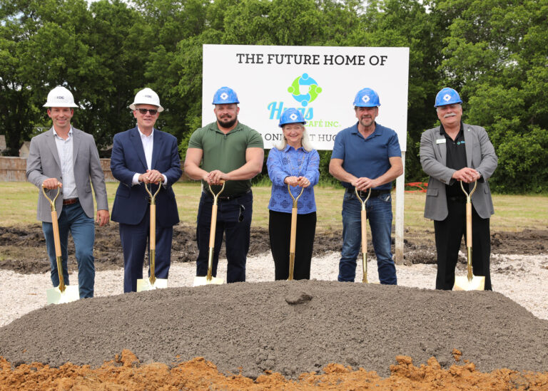 Six people in hard hats stand behind a mound of dirt, holding shovels, at a Dallas groundbreaking.