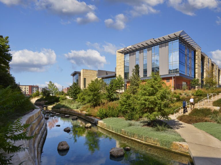 A modern building with large glass windows, built using self perform construction, sits beside a landscaped stream and walking path under a partly cloudy sky. Two people are walking up stairs near the water.