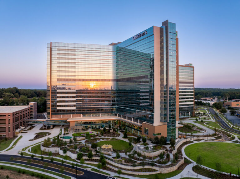 A modern glass-fronted hospital building labeled “Children’s” is shown at sunset, with landscaped grounds and adjacent parking areas.