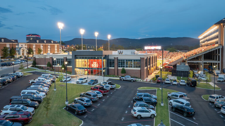A sports complex with a lit stadium packed with people, surrounded by parked cars in the evening and hills in the background, showcases the quality of self perform construction.