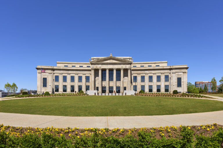 A large, neoclassical stone building with columns, constructed by Vondace Contracting, stands behind a manicured lawn under a clear blue sky.
