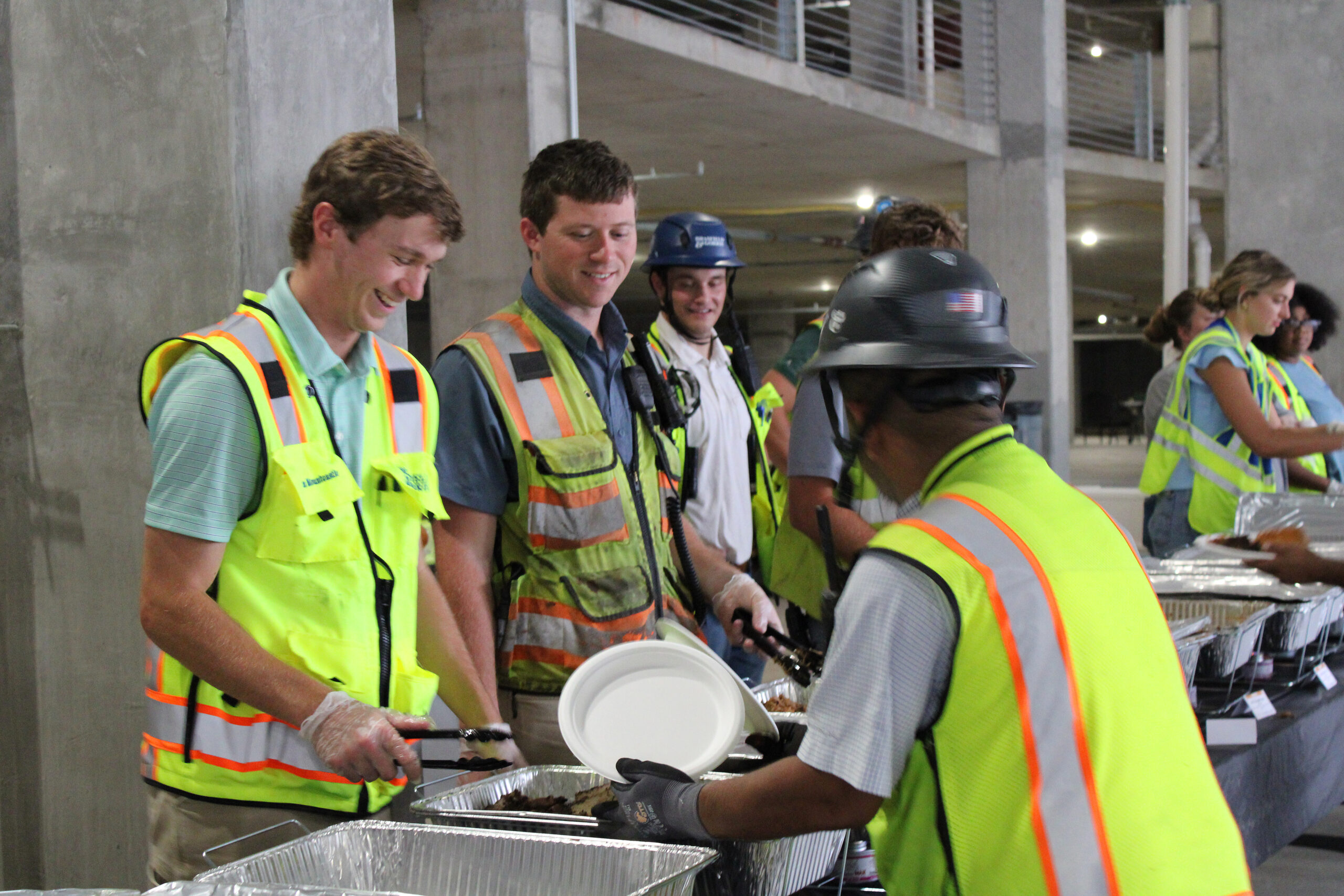 Construction workers in safety vests and helmets serve food onto plates at a buffet-style table in an under-construction setting.