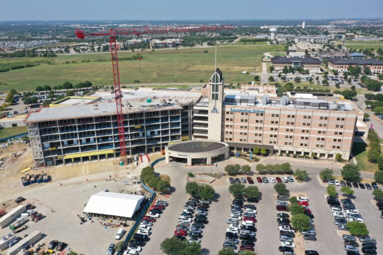 Aerial view of a hospital with a large parking lot, adjacent to a building under construction with a red crane, surrounded by green fields and other buildings.