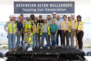 A group of construction workers and staff pose on a stage in front of a banner reading "Ascension Seton Williamson Topping Out Celebration.