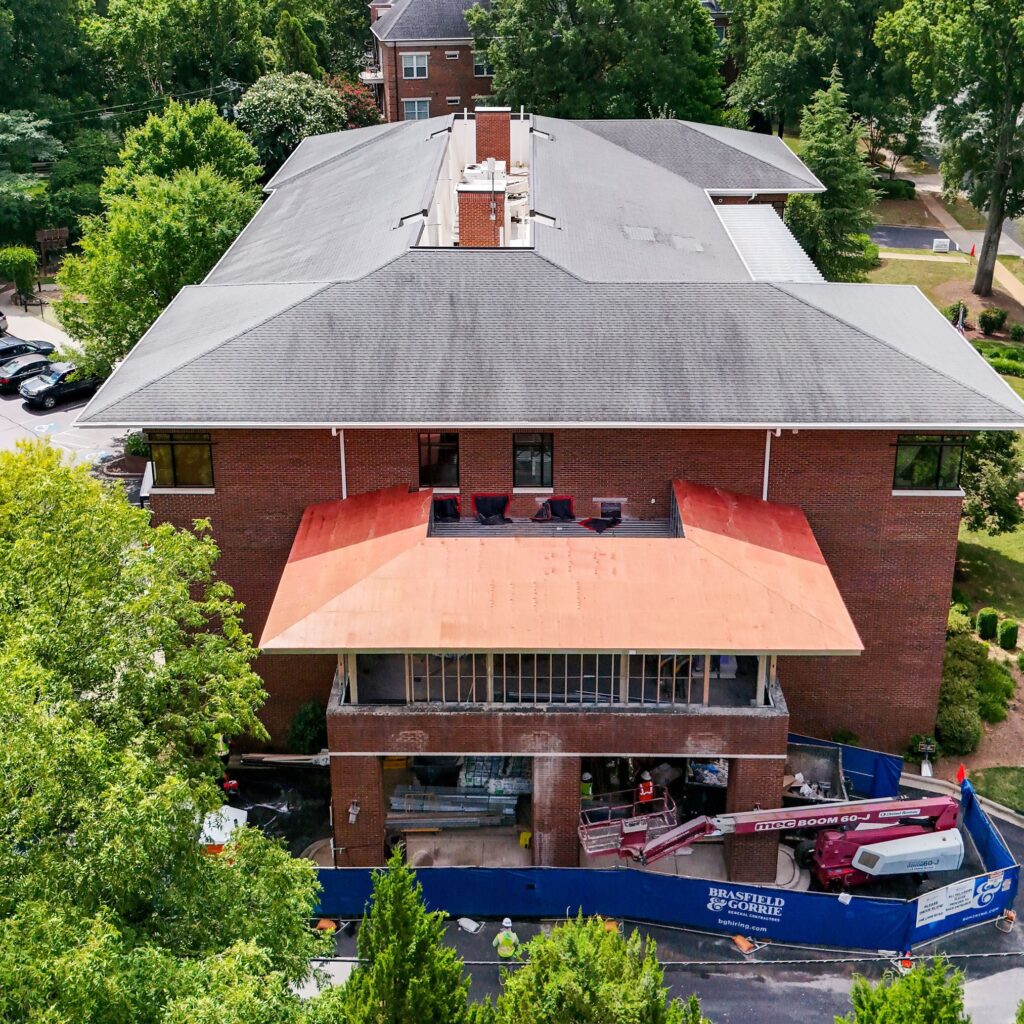 A brick building at a construction milestone, with a partially constructed balcony and equipment in front, surrounded by trees and parked cars—part of the Ronald McDonald House Greater Charlotte project.