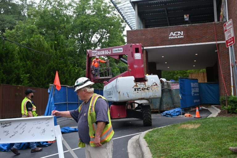 A construction worker signs a white beam near a lift and construction site, celebrating a construction milestone for Ronald McDonald House Charities of Greater Charlotte, while other workers operate machinery and manage the area.