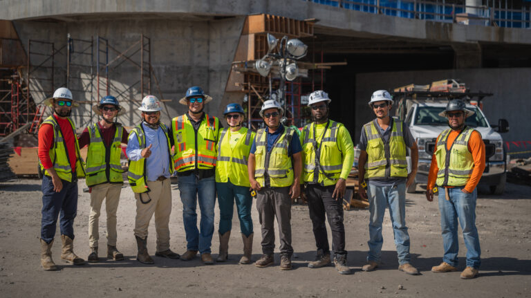 Ten construction workers wearing safety vests and helmets stand in a line at a construction site with equipment and scaffolding visible in the background.