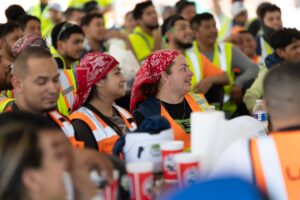 A group of construction workers wearing safety vests and bandanas sit closely together, smiling and listening attentively at a topping out event on a construction site in progress.