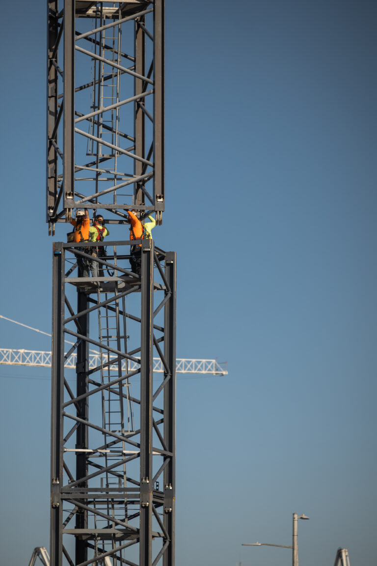 Two construction workers in high-visibility clothing work on a metal structure, showcasing the design build process against a clear blue sky, with cranes visible in the background.