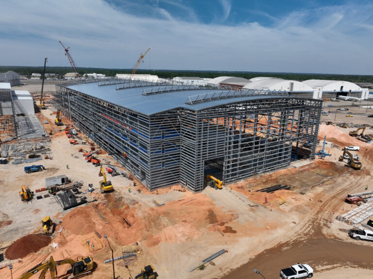 A large metal-frame building is under construction at an industrial site, showcasing a design build approach. The area is surrounded by construction vehicles, equipment, and materials.