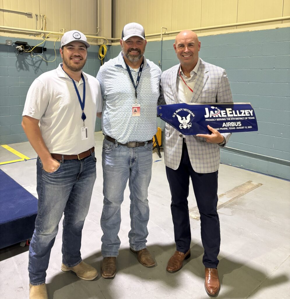 Three men stand indoors; the man on the right holds a blue commemorative propeller blade labeled "Jake Ellzey Airbus," celebrating the Airbus helicopter expansion.