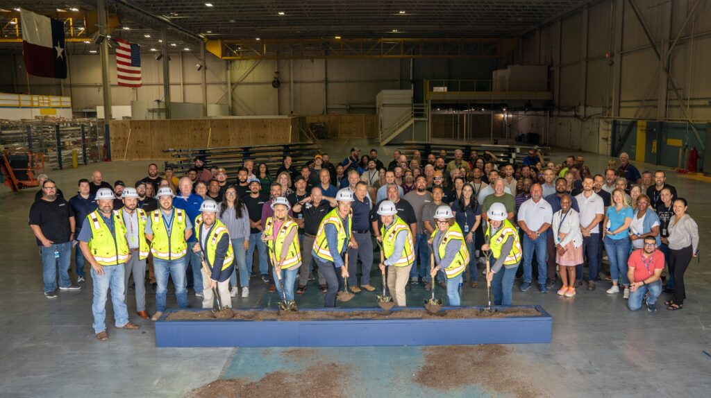 A large group of people, some in construction vests and helmets, stand inside a spacious warehouse for the Airbus helicopter expansion groundbreaking ceremony with Vondace Contracting.