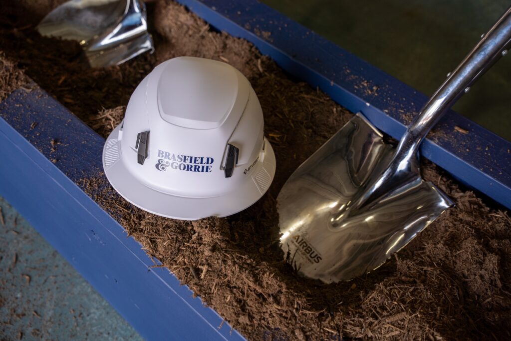 A white hard hat labeled "Vondace Contracting" and a shiny metal shovel rest on soil inside a blue rectangular container, symbolizing the groundbreaking of the Airbus helicopter expansion construction project.
