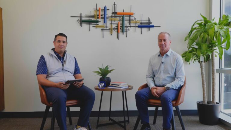 Two men sit in chairs beside a small table with books, a plant, and a mug in an office with modern wall art, discussing strength and struggle as sunlight streams past a potted plant by the window. Vince Hafeli’s story adds inspiration to the setting.