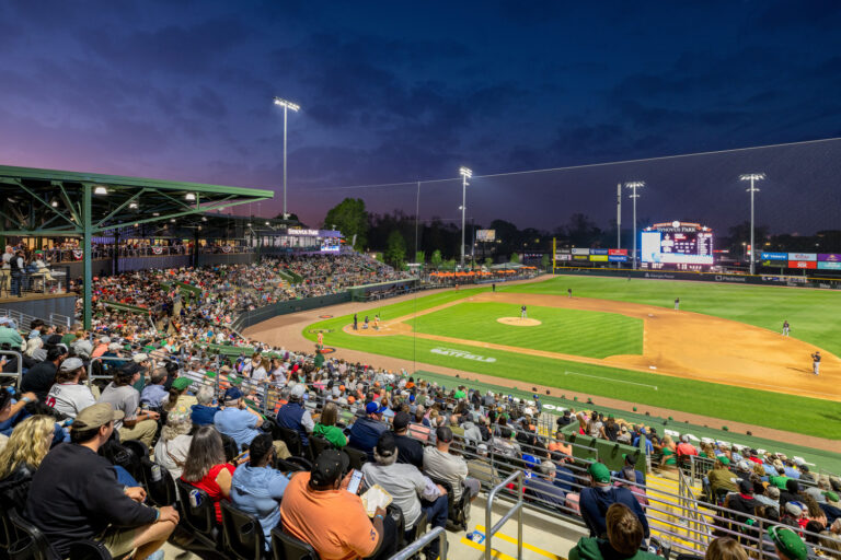 A large crowd watches a Columbus Clingstones baseball game at Synovus Park in Columbus, Georgia, during dusk, with the stadium's bright lights highlighting players on the field, and the scoreboard visible in the background.