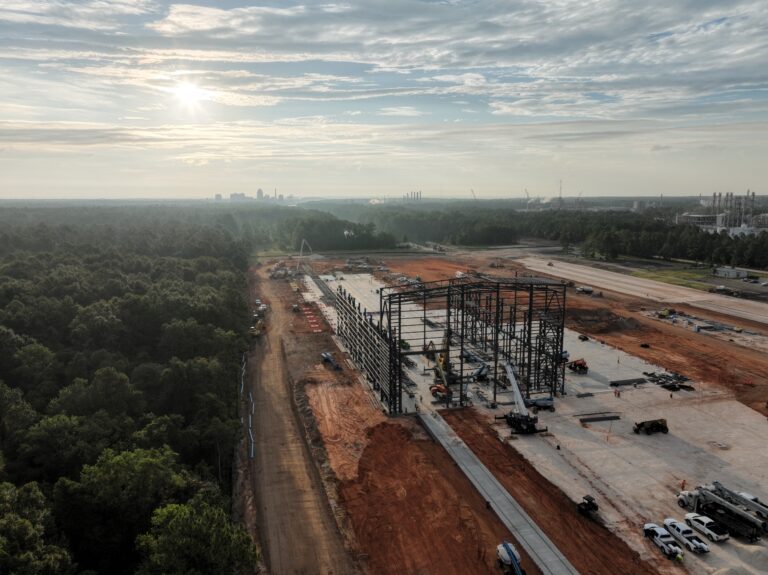 Aerial view of a Vondace Contracting construction site with steel framework rising on a large concrete foundation, bordered by red dirt and forested areas under a partly cloudy sky.