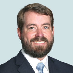 A man with light skin, brown hair, and a beard, wearing a dark suit, white shirt, and light blue tie, smiles in front of a plain light background