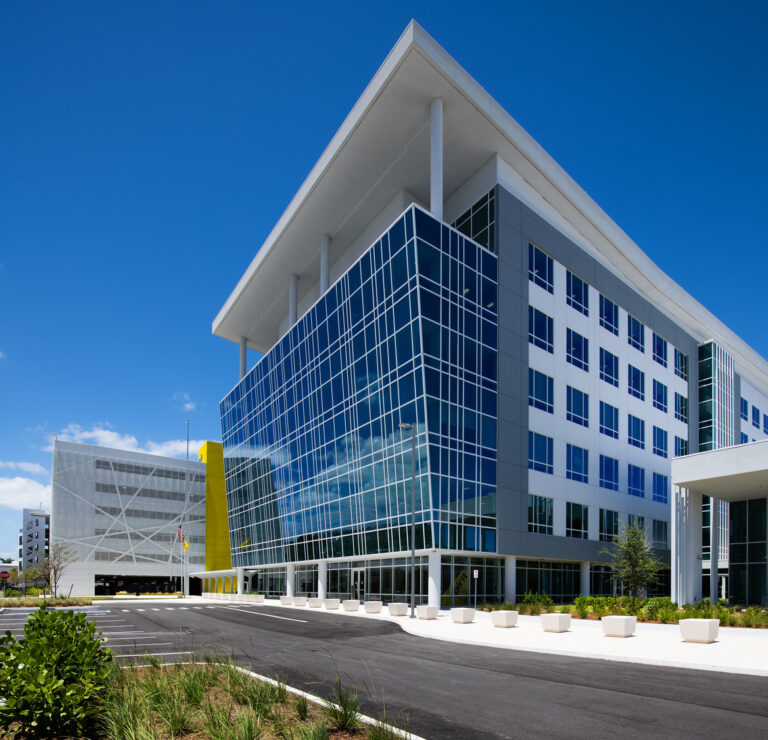 A modern multi-story office building, designed as a headquarters, features large glass windows, white and yellow accents, and a landscaped exterior under a clear blue sky.