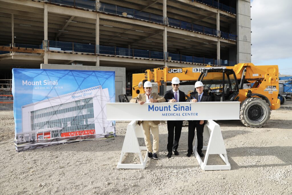 Three people in business attire and hard hats stand at a Miami construction site for Mount Sinai Medical Center, posing with a large white beam and banner in front of an unfinished building and forklift.