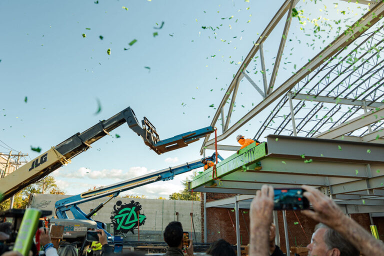 Construction workers secure a steel beam at the future Trueline entertainment venue in Greenville as green confetti falls and people watch and take photos. The milestone is part of Vondace Contracting’s ongoing work on the site.