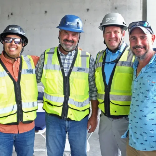 Four men pose together, two wearing construction vests and helmets, standing indoors against a concrete wall.
