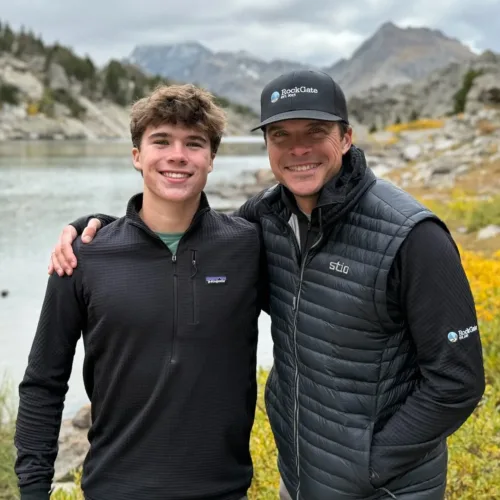 Reed and his son wearing dark jackets stand close together and smile in front of a mountain lake surrounded by rocks and yellow foliage.