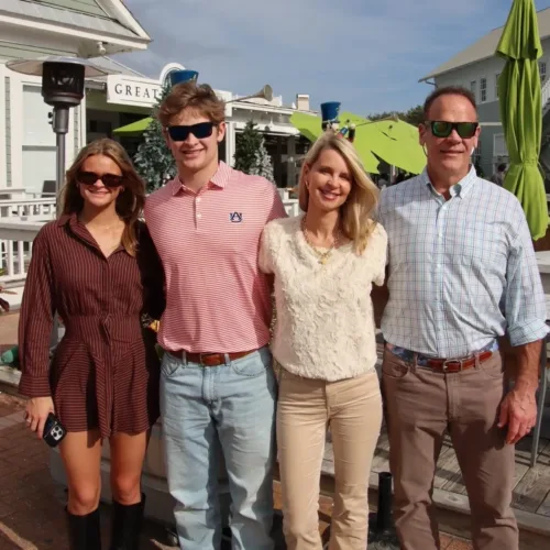 Four adults stand side by side outdoors in sunny weather, wearing casual clothes and sunglasses, with houses and umbrellas in the background.