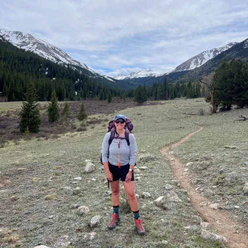 A person wearing hiking gear stands on a trail in a valley surrounded by mountains with snow-capped peaks and pine trees under a cloudy sky.