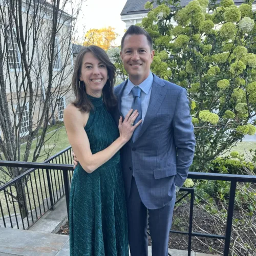 A woman in a green dress and Mike Foushee in a blue suit stand together outside on a stone patio, smiling at the camera. Shrubs and a building are visible in the background.